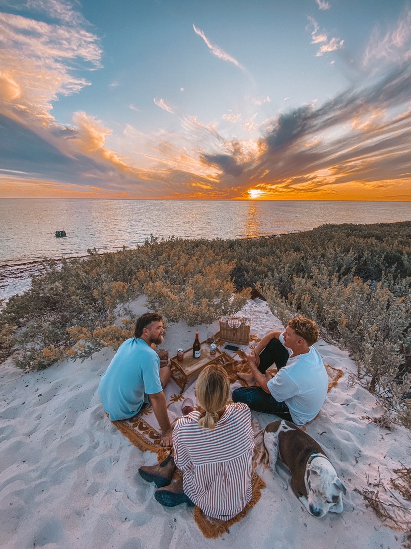 Endless Summer throw rug laid out on the beach, 3 friends are sitting on it enjoying drinks as they watch the sunset over the ocean