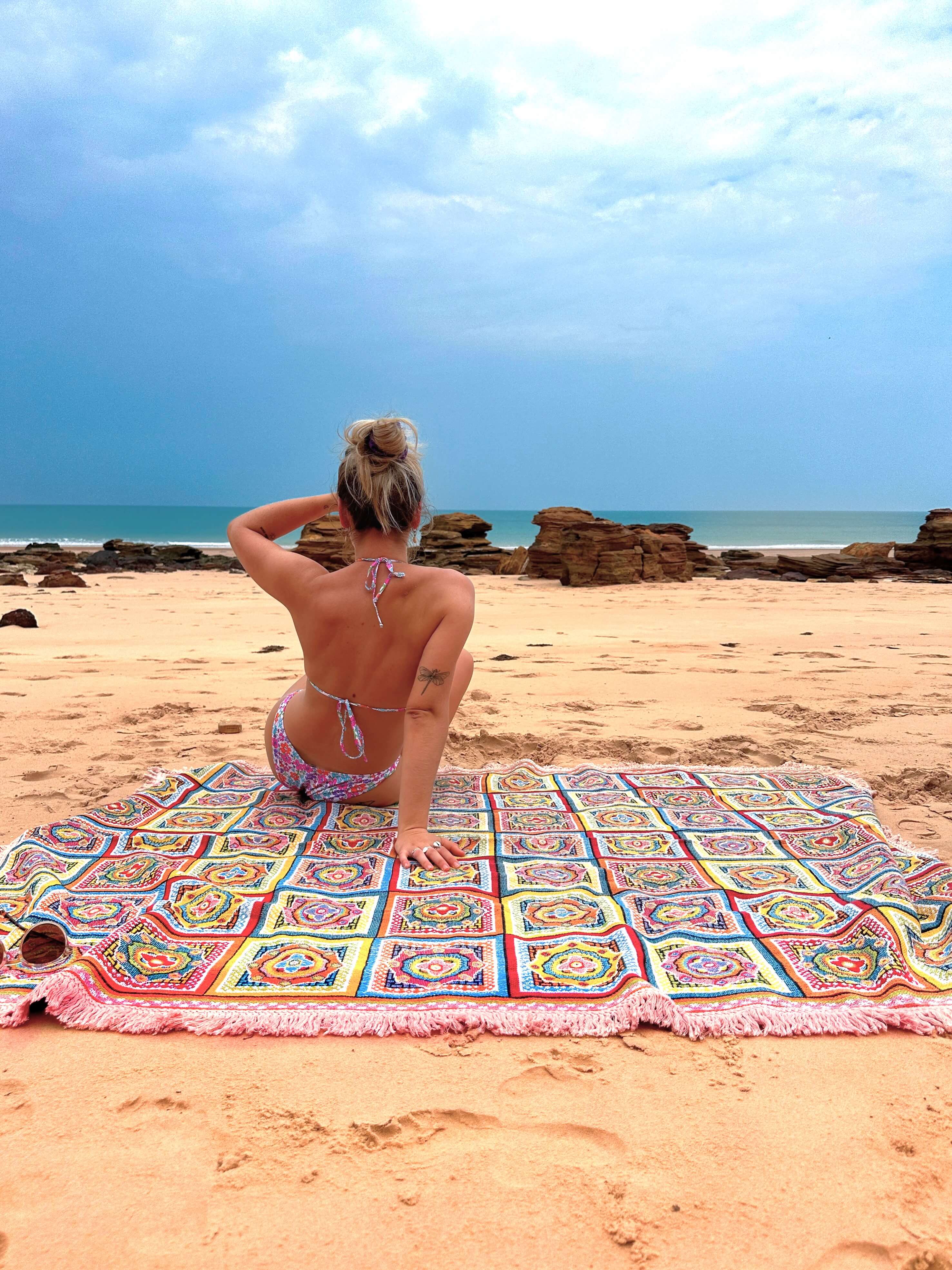 Forever Young throw rug on the sand overlooking a storm rolling in over the ocean. Young woman sits on throw beach blanket looking out.