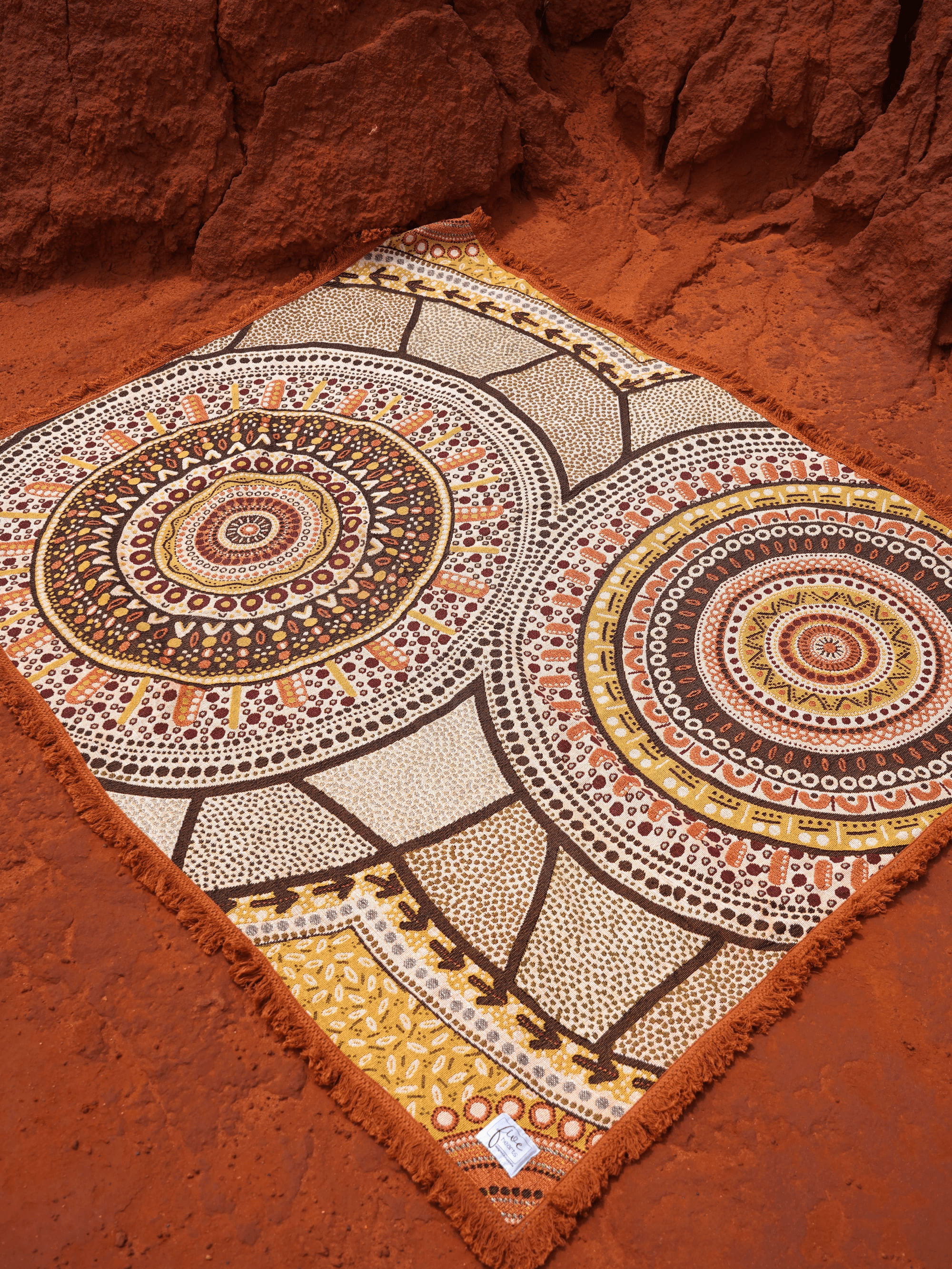 Stomping Ground Throw Rug or Picnic Blanket. Aboriginal art design, Kalkadoon artist. Tones for oranges and browns. Rug laid on red dirt in WA