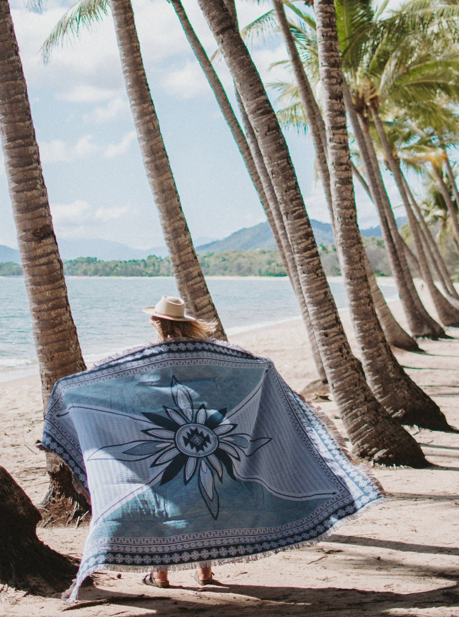 Shooting Stars blue throw rug, picnic blanket around shoulders of woman walking on beach, overlooking ocean in Far North Qld. Designed in Aus.