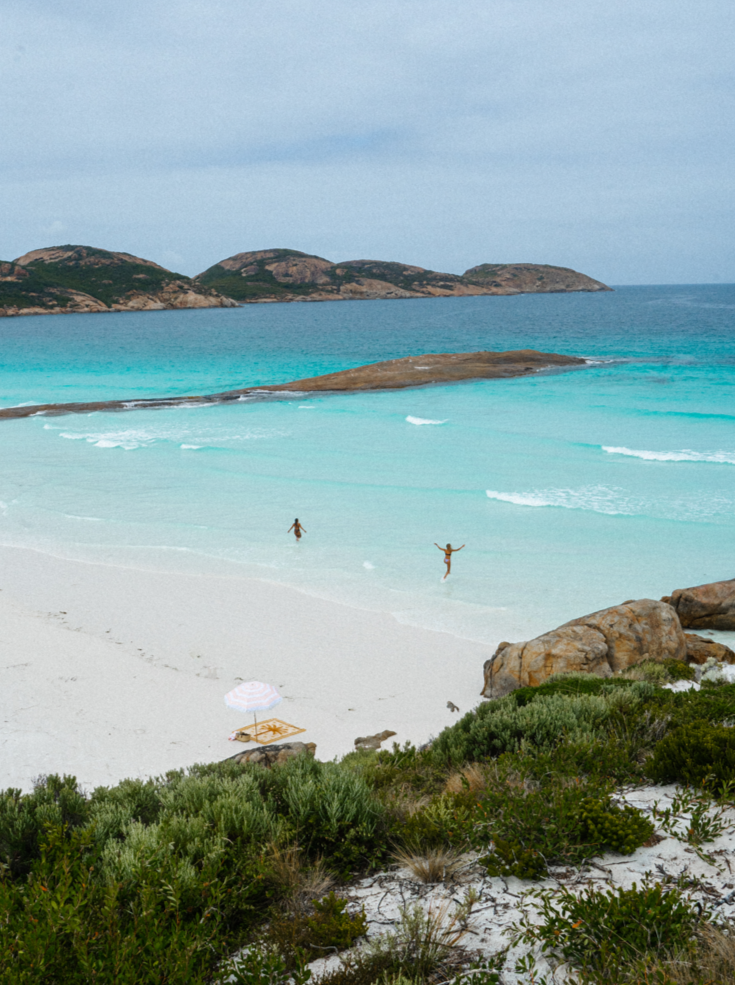 Endless Summer throw blanket on a white sandy beach with a white umbrella over it and two people playing in the ocean behind