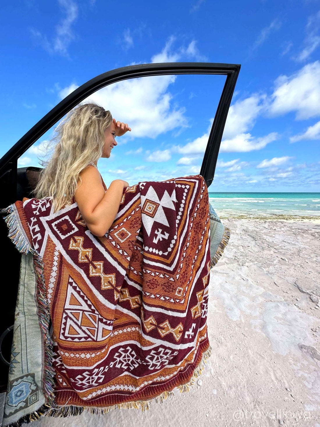 Desert Night throw rug and picnic blanket laying on beach, crystal blue water in background. Shades of brown, boho rug, must-have van life accessory. Designed in Australia for adventure.