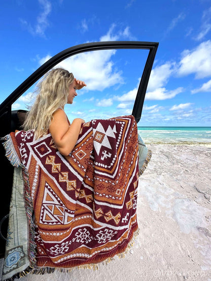 Desert Night throw rug and picnic blanket laying on beach, crystal blue water in background. Shades of brown, boho rug, must-have van life accessory. Designed in Australia for adventure.