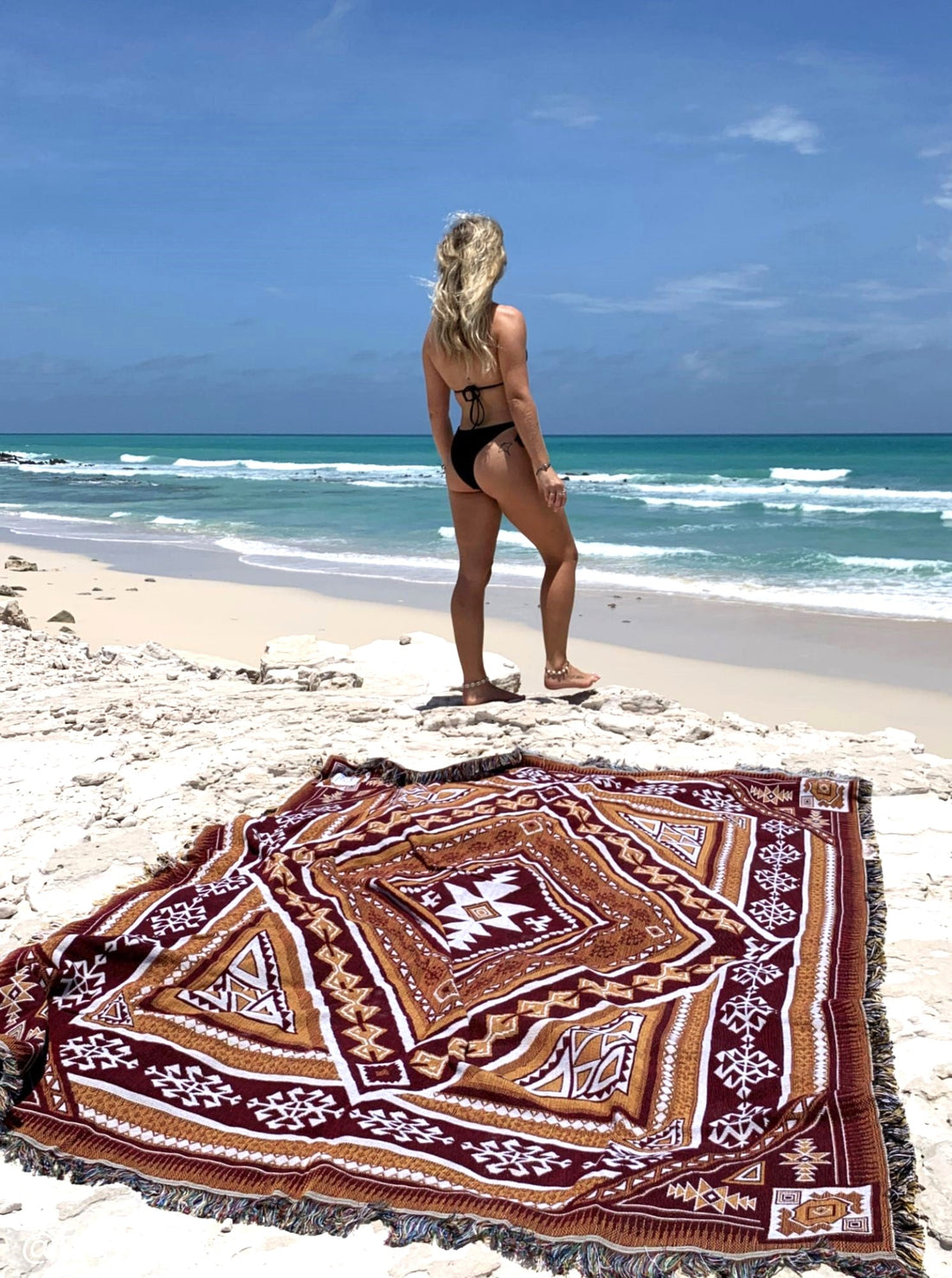 Desert Night throw rug and picnic blanket laying on beach, crystal blue water in background. Shades of brown, boho rug, must-have van life accessory. Designed in Australia for adventure.