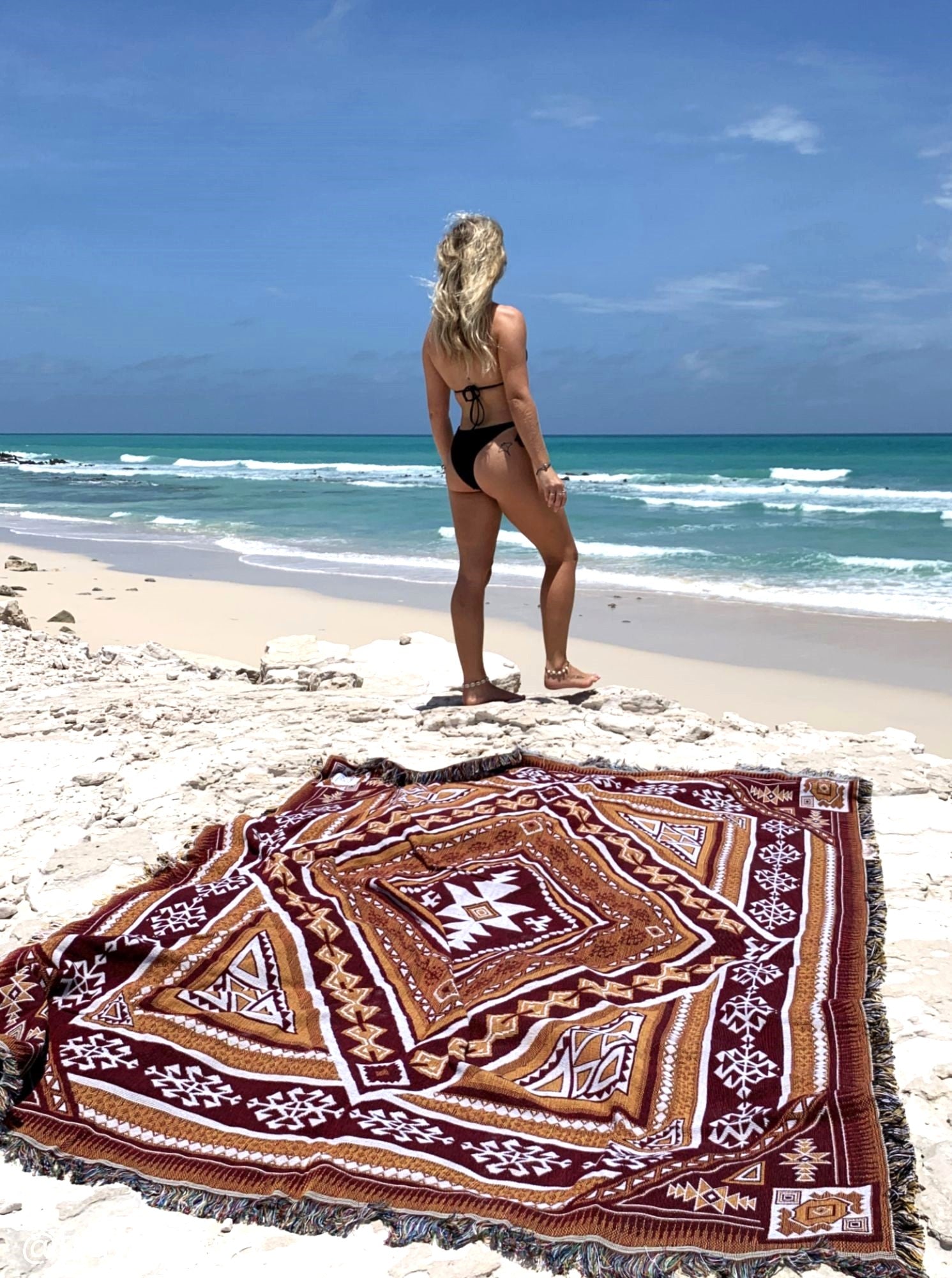 Desert Night throw rug and picnic blanket laying on beach, crystal blue water in background. Shades of brown, boho rug, must-have van life accessory. Designed in Australia for adventure.